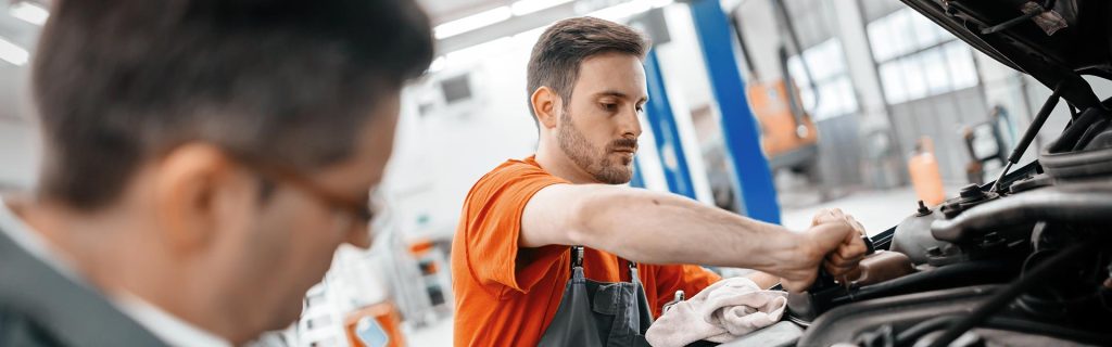 A mechanic working on a car's engine while another person observes.